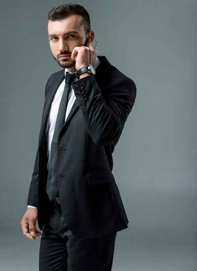 Handsome security guard listening message with security earpiece and looking away isolated on grey